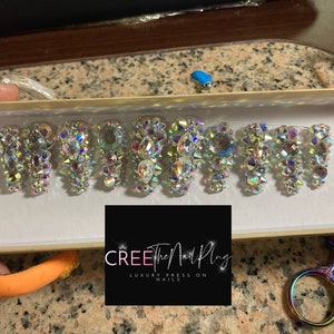 May include: A set of ten press-on nails with a clear base and iridescent rhinestones. The nails are arranged in a box with the brand name "CREE the Nail Plug" and the text "Luxury Press On Nails".