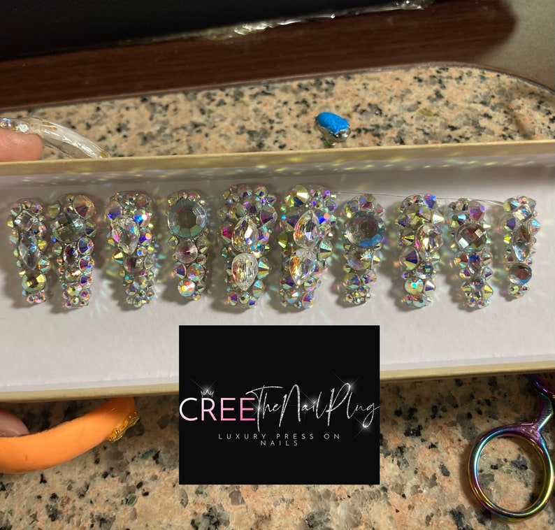 May include: A set of ten press-on nails with a clear base and iridescent rhinestones. The nails are arranged in a box with the brand name "CREE the Nail Plug" and the text "Luxury Press On Nails".