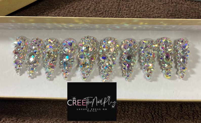 May include: Ten clear press-on nails with iridescent rhinestones. The nails are arranged in a row in a white box. The box has a black label with the text "CREE The Nail Plug LUXURY PRESS ON NAILS".