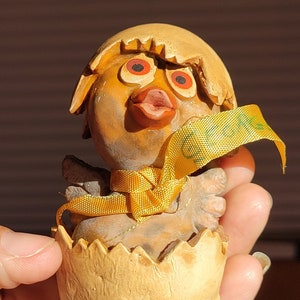 Puede incluir: Figura de madera de un pollito saliendo de un huevo. El pollito tiene patas naranjas, una cinta amarilla y ojos con anillos rojos. La cáscara del huevo es de color marrón claro con bordes dentados. La figura se sostiene en una mano.