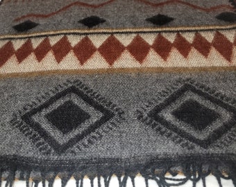 May include: A rectangular scarf with a geometric pattern in shades of gray, brown, and black. The design includes diamond and zigzag shapes. The scarf has a fringed edge.