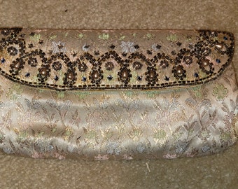 May include: A beige satin clutch with a floral pattern. The flap is embellished with beads and pearls in shades of brown, black, and blue. The clutch is a rectangular shape and appears to be closed.