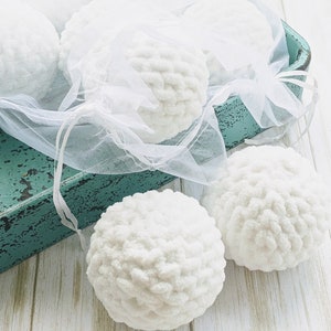 May include: A collection of white, fluffy, crocheted spheres. Some are presented within a sheer white bag, secured with a ribbon. Others are arranged on a distressed teal tray and a light wood surface. The spheres have a soft, textured appearance.