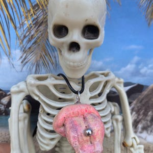 May include: A pink tongue pendant necklace with a silver ring and a silver bead. The necklace is worn by a skeleton.