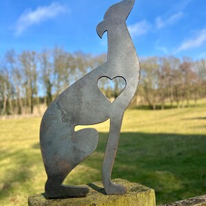 A metal fence topper in the shape of a greyhound/whippet with a heart cut out, placed on a fence with a field and trees in the background.