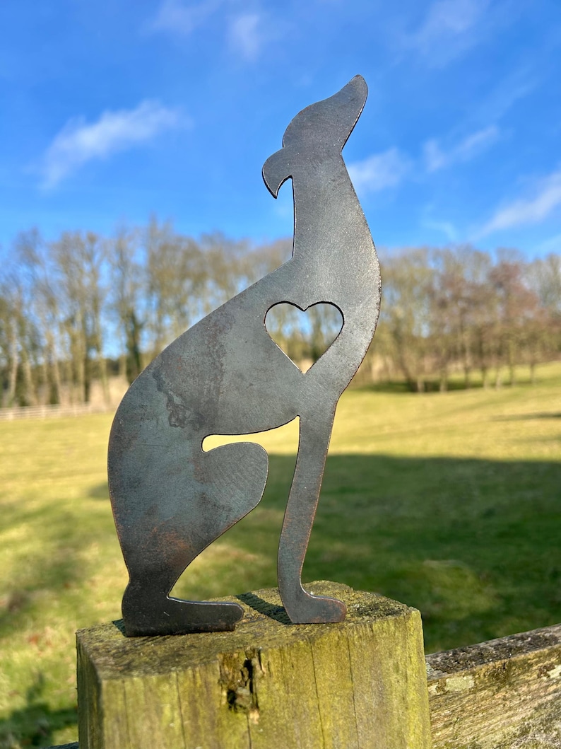 A metal fence topper in the shape of a greyhound/whippet with a heart cut out, placed on a fence with a field and trees in the background.