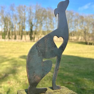 A metal fence topper in the shape of a greyhound/whippet with a heart cut out, placed on a fence with a field and trees in the background.