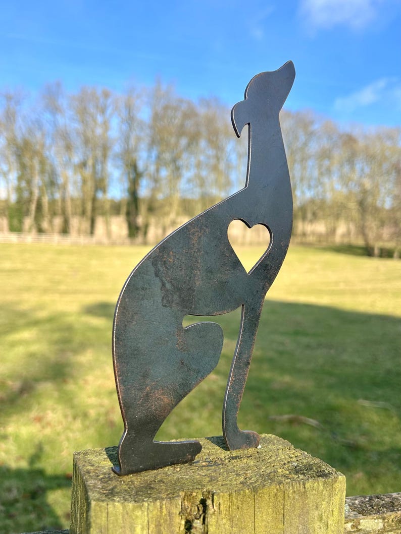 A metal fence topper in the shape of a greyhound/whippet with a heart cut out, placed on a fence with a field and trees in the background.