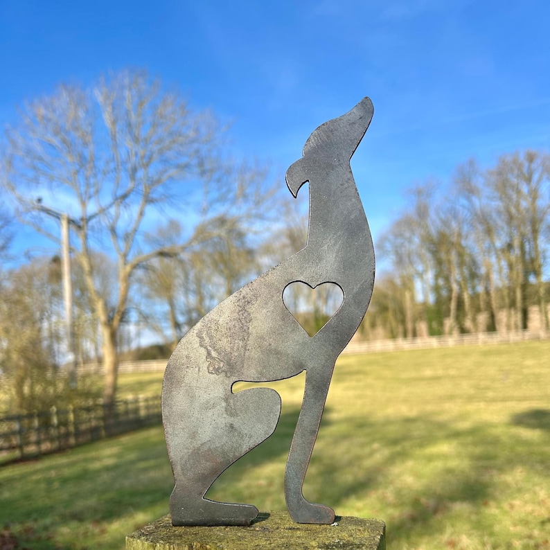 A metal fence topper in the shape of a greyhound/whippet with a heart cut out, placed on a fence with a field and trees in the background.