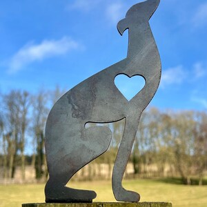 A metal fence topper in the shape of a greyhound/whippet with a heart cut out, placed on a fence with a field and trees in the background.