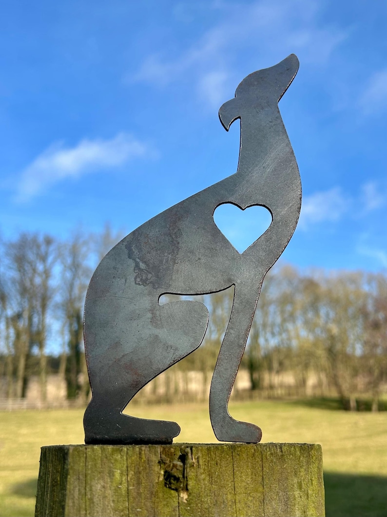 A metal fence topper in the shape of a greyhound/whippet with a heart cut out, placed on a fence with a field and trees in the background.