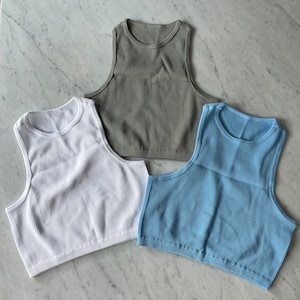 May include: Three ribbed tank tops in different colors: light blue, white, and olive green. The tank tops are sleeveless and cropped.