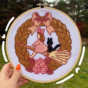 Op de afbeelding: Kruissteekborduurwerk in een houten hoepel. Het ontwerp toont een meisje op een bezem met een zwarte kat, omringd door een krans en strikken. Het kleurenpalet omvat roze, bruin en zwart, wat een grillige en handgemaakte esthetiek creëert.