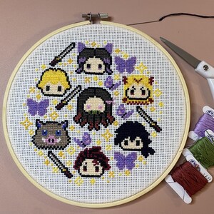 May include: A completed cross-stitch project featuring cartoon character portraits, swords, butterflies, and starbursts. The design is set within a wooden embroidery hoop. Additional craft supplies include scissors and spools of thread.