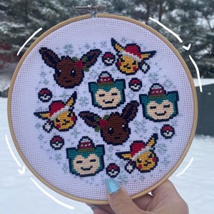 May include: A cross-stitch embroidery hoop featuring various Pokémon characters in festive holiday attire. The design includes Eevee, Pikachu, and Snorlax, each wearing a Santa hat. The background is white with small snowflake accents, all within a wooden hoop.