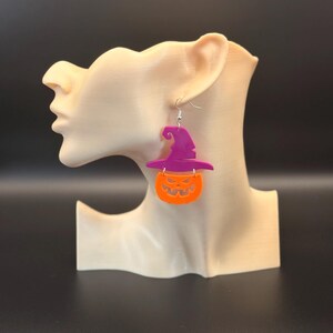 May include: Halloween-themed earrings featuring a purple witch hat and an orange jack-o'-lantern design. The earrings are suspended from silver-colored hooks and are displayed on a mannequin head. The earrings are a festive accessory for the Halloween season.