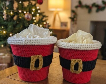 May include: Two cylindrical crochet baskets designed to resemble Santa Claus suits. Each basket features a red body, a black belt with a gold-colored buckle, and a white crocheted rim. Both containers are filled with light-colored tissue paper.