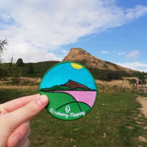 May include: A round, green, pink, and blue embroidered patch with a mountain scene and the text "Roseberry Topping".