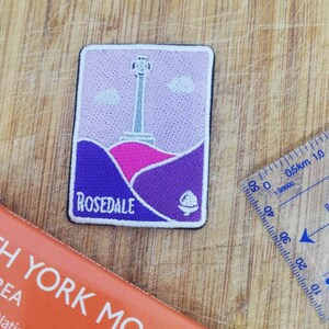 Peut inclure: Un patch brodé avec un motif violet, rose et blanc. Le patch représente une tour avec un toit blanc et une colline rose en arrière-plan. Le texte "ROSEDALE" est imprimé en blanc sur le patch.