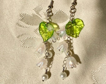 May include: A pair of dangle earrings with white flower charms and green leaf charms. The earrings are made with a silver chain and have pearl accents.