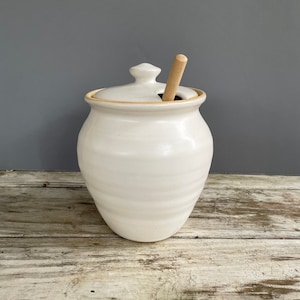 May include: White ceramic honey pot with a wooden honey dipper. The pot has a lid and a slightly rounded shape.
