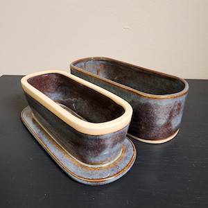 May include: Two ceramic oval-shaped dishes with a blue and brown glaze. The smaller dish has a cream-colored rim and sits on top of a larger dish.