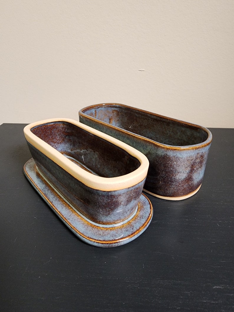May include: Two ceramic oval-shaped dishes with a blue and brown glaze. The smaller dish has a cream-colored rim and sits on top of a larger dish.