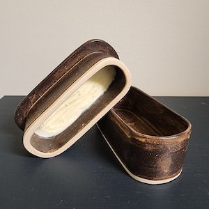 May include: A brown ceramic butter dish with a lid. The dish is oval shaped and has a white rim. The lid is slightly ajar, revealing a dollop of butter inside.