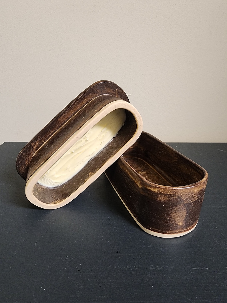 May include: A brown ceramic butter dish with a lid. The dish is oval shaped and has a white rim. The lid is slightly ajar, revealing a dollop of butter inside.