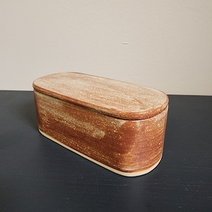 May include: A rectangular ceramic butter dish with a lid. The dish is a light brown color with a speckled glaze. The lid is slightly lighter in color and has a smooth finish.