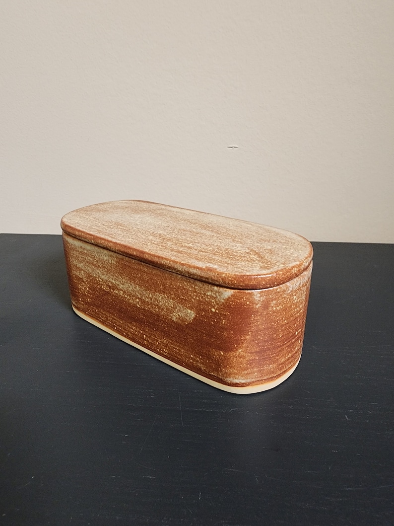 May include: A rectangular ceramic butter dish with a lid. The dish is a light brown color with a speckled glaze. The lid is slightly lighter in color and has a smooth finish.