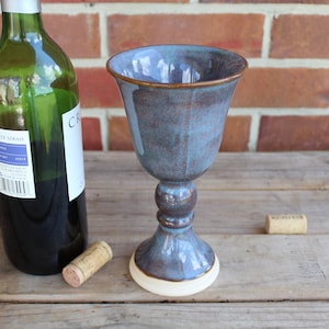 May include: A blue ceramic wine goblet with a white base. The goblet is sitting on a wooden surface next to a bottle of wine and two corks.