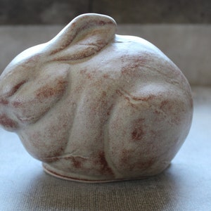 May include: A ceramic rabbit sculpture with a white glaze and brown accents. The rabbit is curled up in a sleeping position.