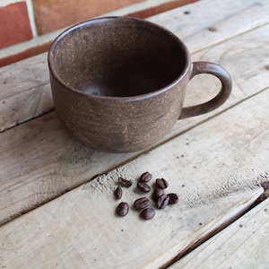 May include: A brown ceramic mug with a handle sits on a wooden surface with a scattering of coffee beans.
