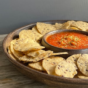 May include: A brown ceramic serving dish with a small bowl in the center filled with red salsa. The dish is filled with a ring of light brown tortilla chips.