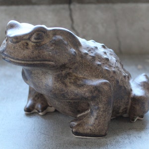 May include: A brown ceramic toad figurine with a textured surface. The toad is sitting on a light gray surface.