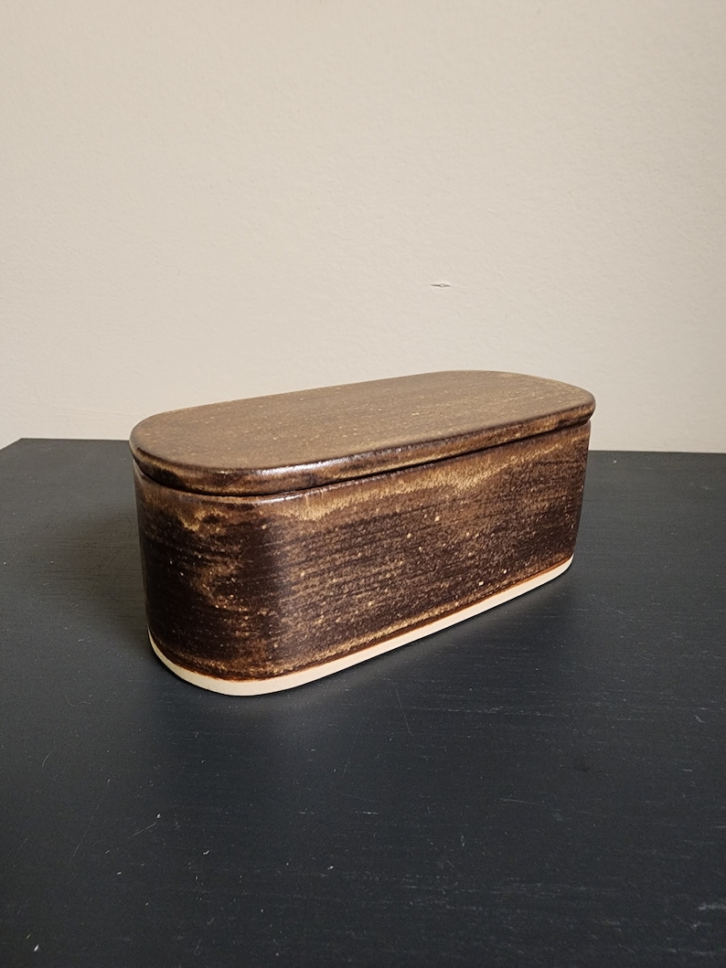 May include: A brown ceramic butter dish with a lid. The dish has a slightly oval shape and a white rim.