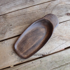 May include: A brown ceramic serving dish with a slightly rounded shape and a small handle on one end. The dish has a speckled, rustic appearance.
