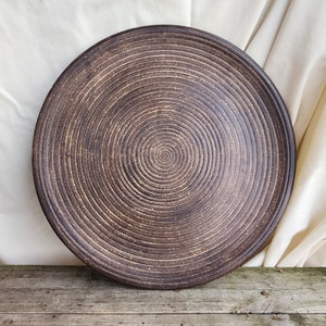 May include: A large, round, brown ceramic plate with a concentric circle design. The plate has a rough, textured surface.