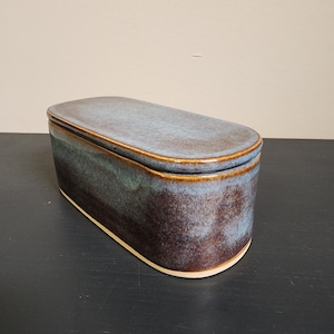 May include: A ceramic butter dish with a lid. The dish is oval shaped and has a blue and brown glaze. The lid is slightly domed and has a light blue glaze.