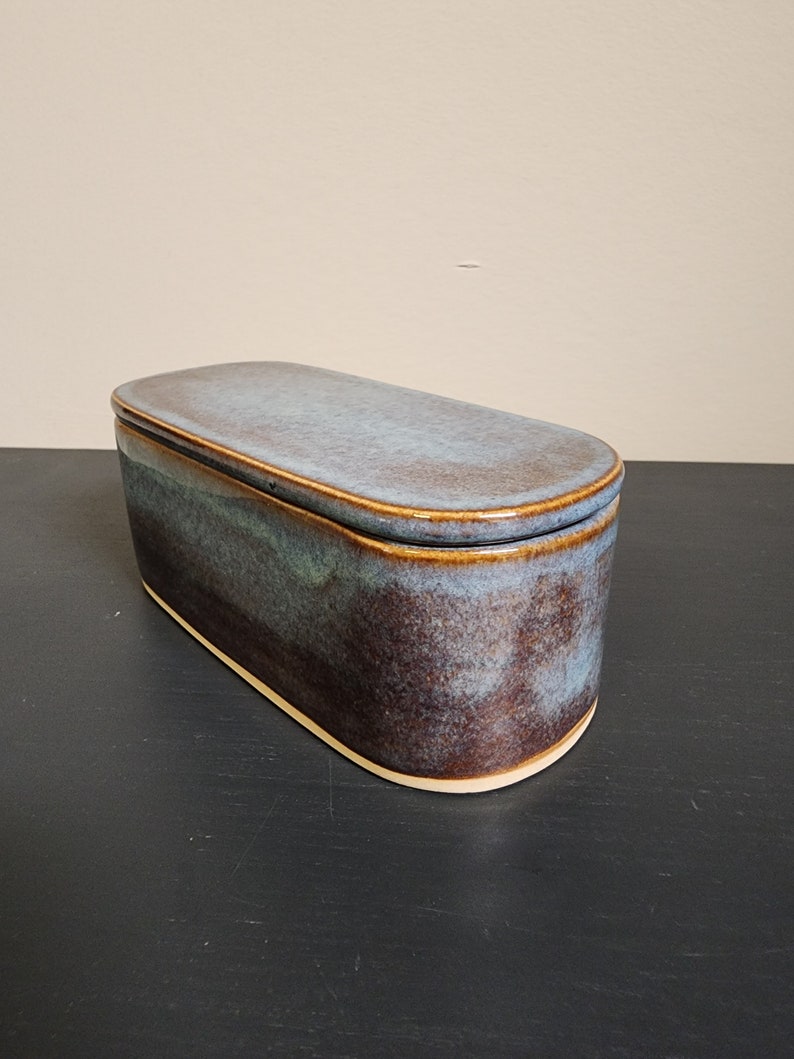 May include: A ceramic butter dish with a lid. The dish is oval shaped and has a blue and brown glaze. The lid is slightly domed and has a light blue glaze.