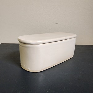 May include: A white ceramic butter dish with a lid. The dish is oval shaped and has a smooth, glossy finish.