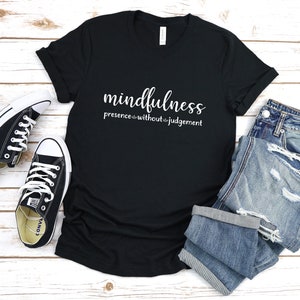 以下が含まれることがあります： 白い文字で「mindfulness presence without judgement」と書かれた黒いTシャツ。白い木製の表面に、黒いコンバースのスニーカーと青いジーンズと一緒に置かれています。