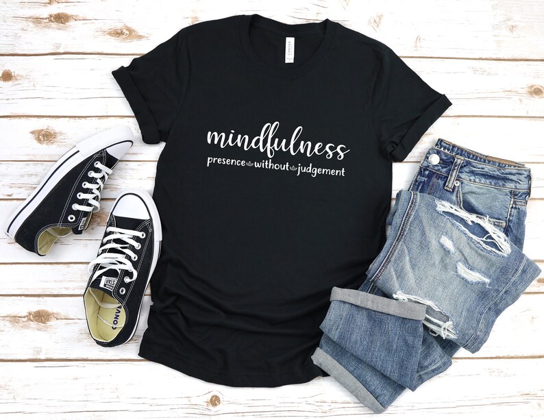 以下が含まれることがあります： 白い文字で「mindfulness presence without judgement」と書かれた黒いTシャツ。白い木製の表面に、黒いコンバースのスニーカーと青いジーンズと一緒に置かれています。