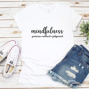 以下が含まれることがあります： 黒で「mindfulness presence without judgement」とプリントされた白いTシャツ