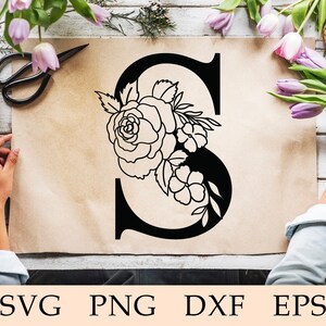 May include: A black and white floral design featuring a large rose and smaller flowers arranged around the letter S. The design is on a brown paper background.