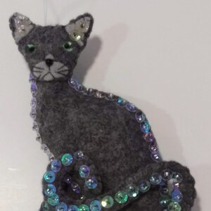 May include: A gray cat ornament with green eyes and a white muzzle. The cat is embellished with iridescent sequins along its body and tail. The ornament is made of felt and has a string for hanging.