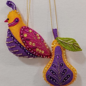 May include: Two handmade felt ornaments: a yellow and purple bird and a purple pear. The bird has pink and purple beaded accents. The pear has a green leaf and gold beaded accents. Both have gold hanging strings.