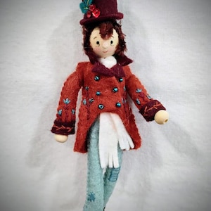 May include: A handmade doll ornament featuring a figure in a red coat with blue accents, light blue pants, and a burgundy top hat. The doll has a wooden head and hands, with brown hair and a white scarf. The ornament is designed for holiday decor.