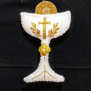 May include: A white chalice ornament with gold accents. The chalice features a gold cross and wheat design, with gold sequins and a gold disc on top. A gold string is attached for hanging.